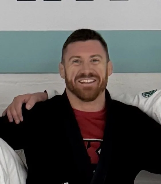 Smiling man with short dark hair and beard, wearing a black jacket over a red shirt, sitting in a hospital bed with teal and white walls in the background.
