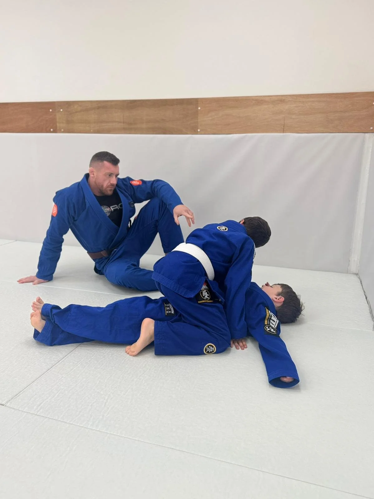 Two men practicing Brazilian Jiu-Jitsu on a mat. One man is on top, pinning the other, who is on his back. Both are wearing blue gis.
