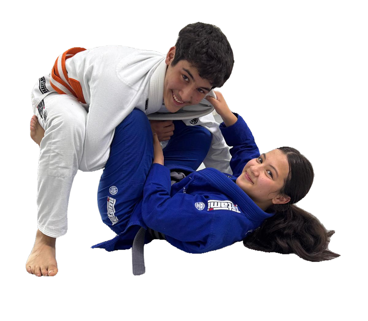 Two people practicing Brazilian Jiu-Jitsu, a man in a white gi and a woman in a blue gi, engaging in a grappling move on the mat. Both are smiling and looking at the camera.