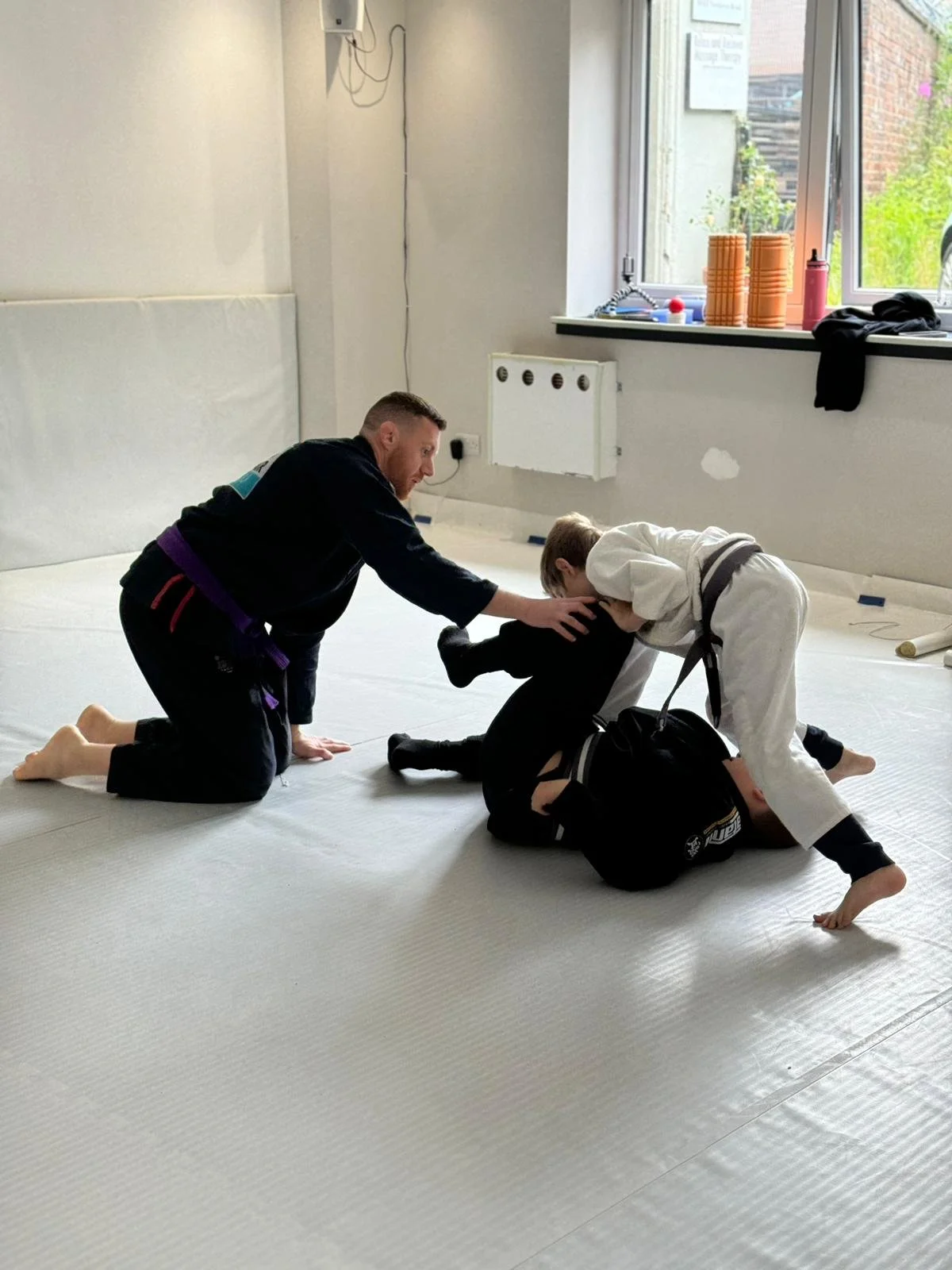 Two children practicing Brazilian Jiu-Jitsu on a mat in a gym, with an instructor assisting each child in gripping and positioning.