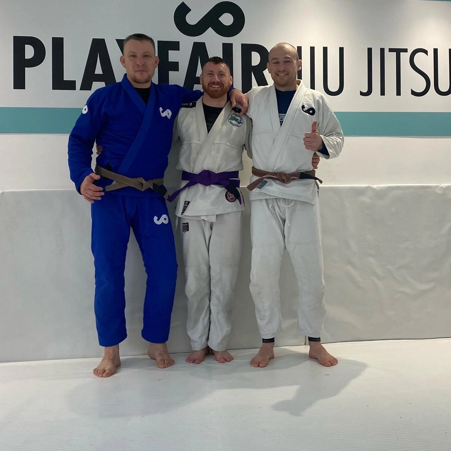 Three men in Brazilian Jiu-Jitsu gis standing together in a dojo, smiling, with one giving a thumbs-up. They are in front of a wall with a sign that reads 'PlayFair BJJ Jiu Jitsu.'