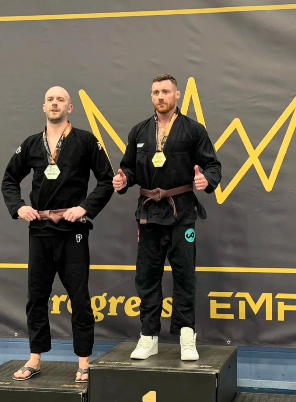 Two men in black martial arts uniforms standing on a winners' podium, holding medals, with a black and yellow backdrop featuring a stylized graph and the text 'rogres' and 'ÊM'. one man gives a thumbs up, the other looks at the camera.