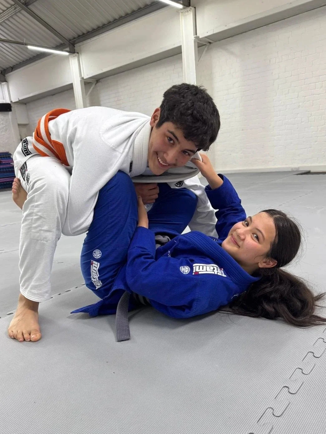 Two Brazilian Jiu-Jitsu practitioners practicing in a gym, with the female in a blue gi on the mat and the male in a white gi on top, both smiling.