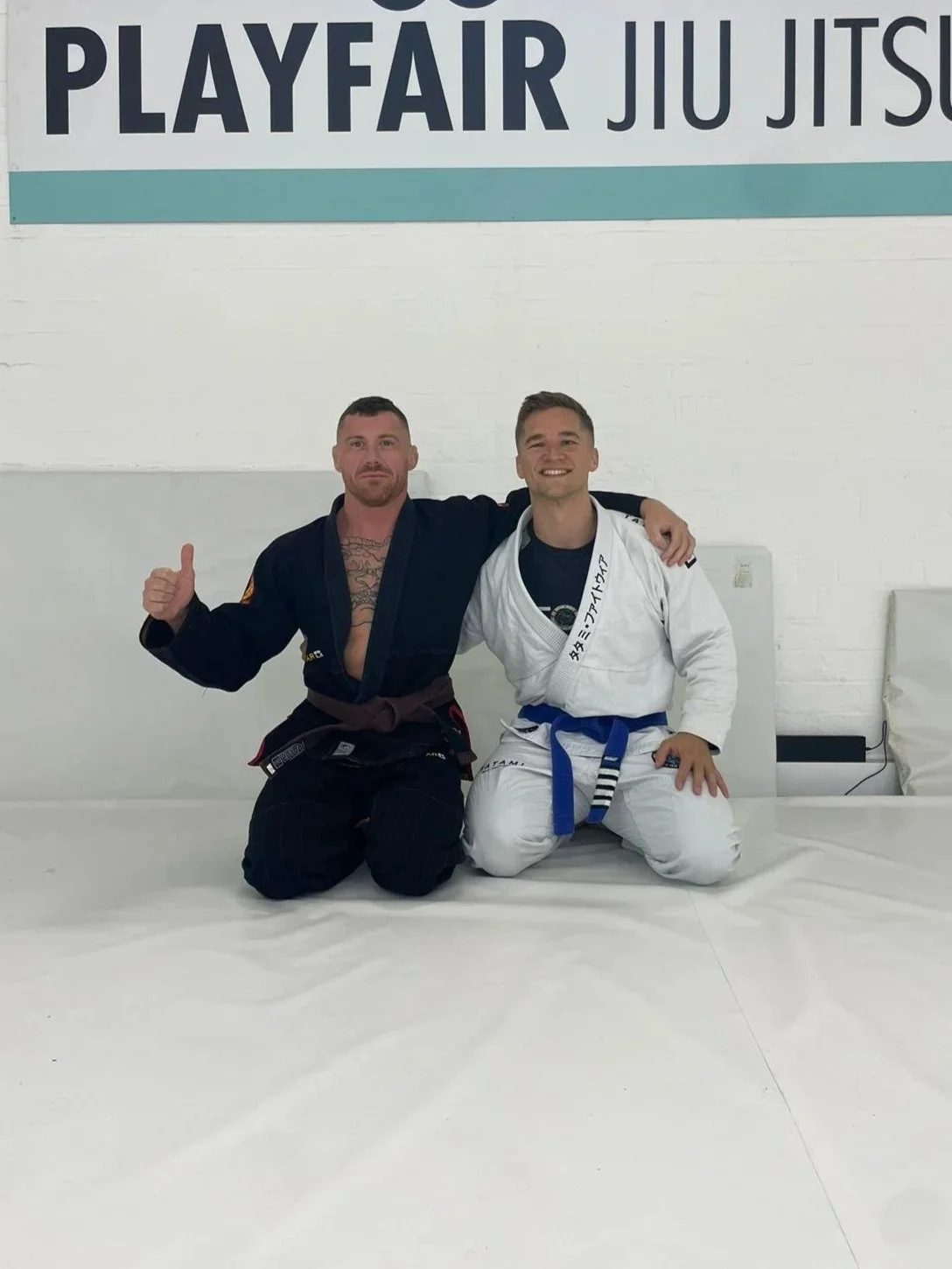 Two men in Brazilian Jiu-Jitsu gis sitting on a padded training mat in a dojo, smiling with arms around each other, under a sign that says 'PlayFair Jiu Jitsu'.