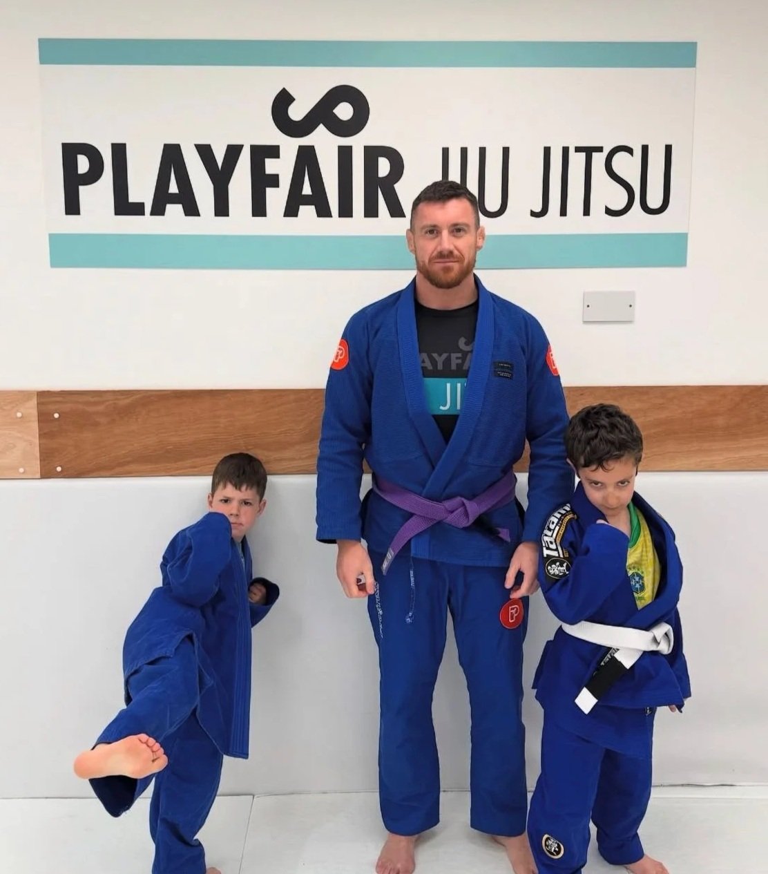 Three people in Brazilian Jiu Jitsu uniforms standing in front of a La Jiu Jitsu sign at a gym.