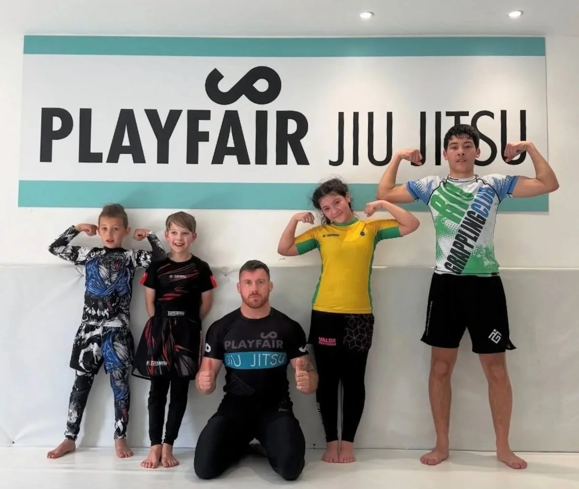 Children and a coach at a Brazilian Jiu-Jitsu gym posing with flexed arm muscles in front of a wall with a PlayFair Jiu-Jitsu sign.