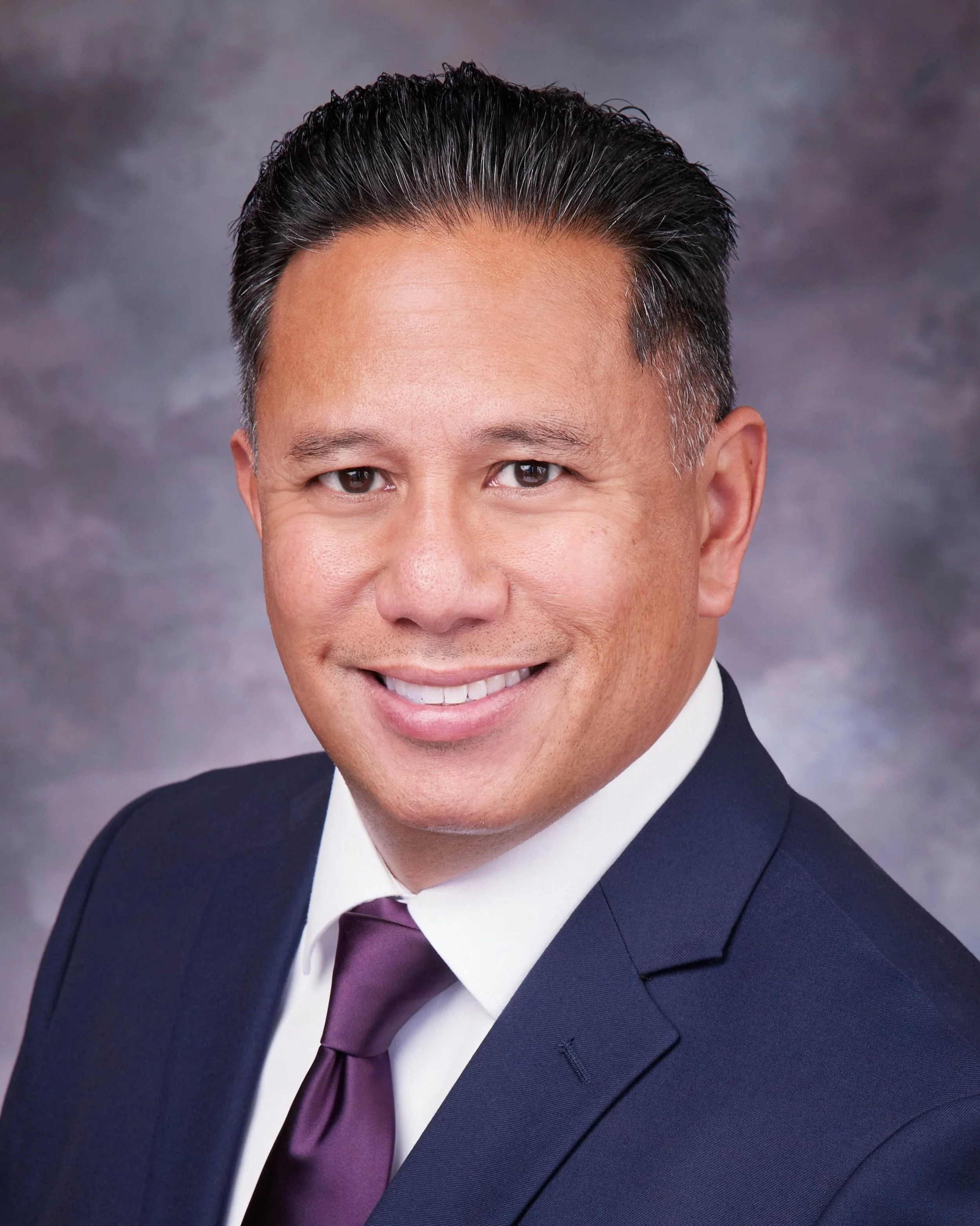 A professional headshot of a man in a dark suit, white shirt, and purple tie, smiling at the camera with a blurred grey background.