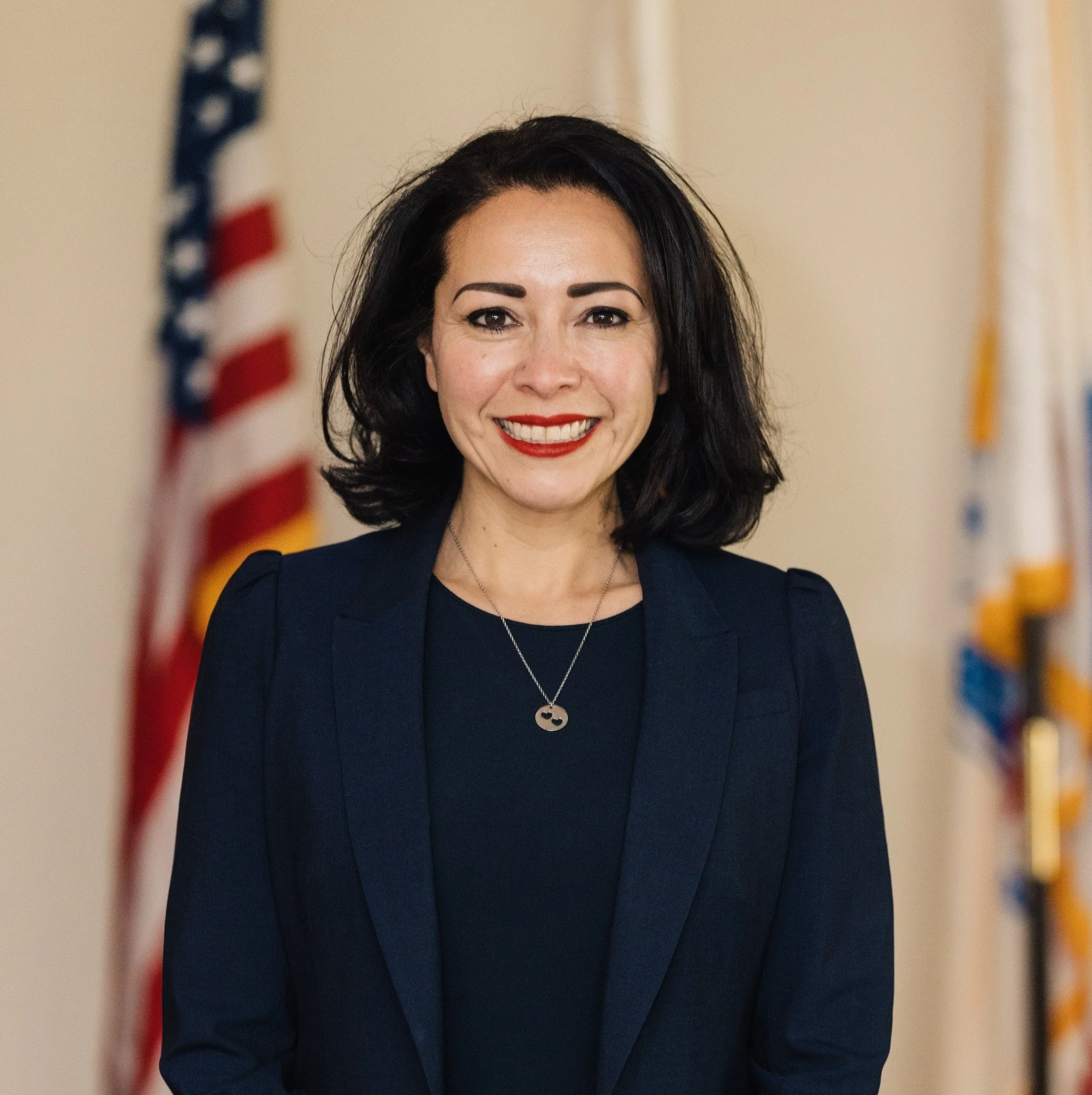 A smiling woman with black hair wearing a navy blazer and a necklace, standing in front of two American flags.