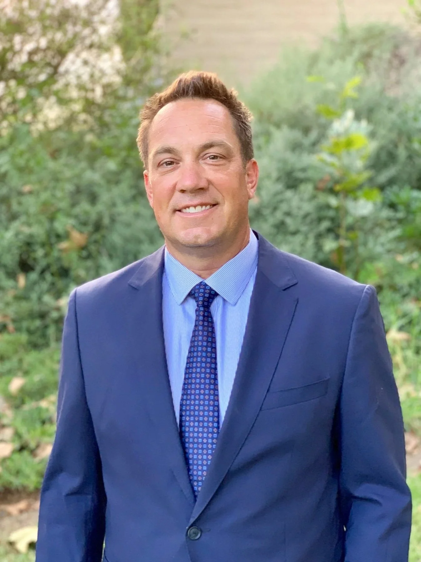 A man with brown hair and a light complexion, wearing a navy blue suit, light blue shirt, and patterned blue tie, smiling outdoors with green trees in the background.
