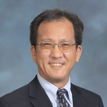 A smiling man wearing glasses, a dark suit, a light shirt, and a patterned tie, posing against a blue background.