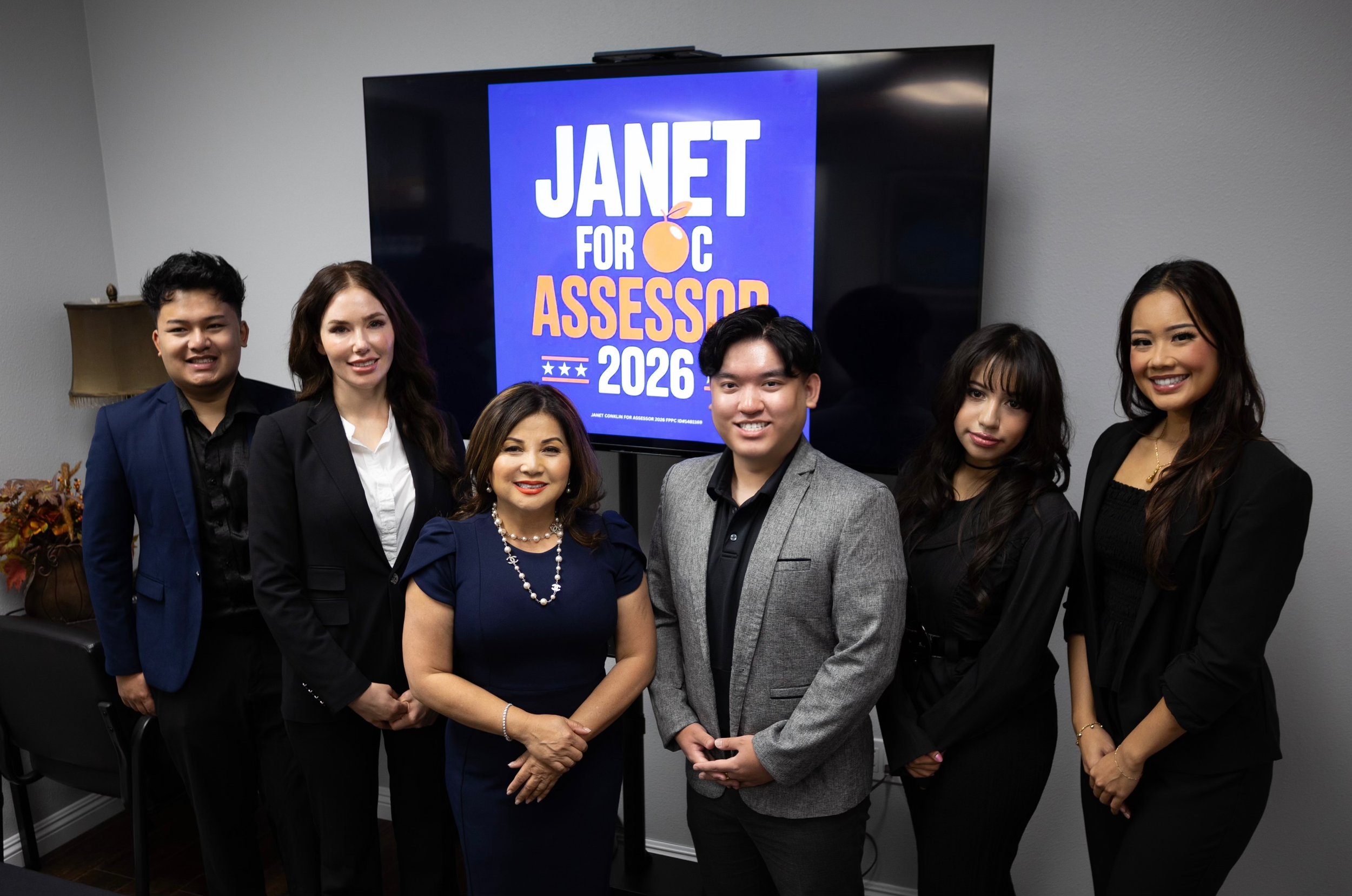 Group of six people: Ethan Tran, Ashley Eagle, Janet Keo Conklin, Cyrus Vong, Katie Ventura, and Marilyn Tap, standing in front of a television screen displaying a graphic for Janet for OC assessor 2026
