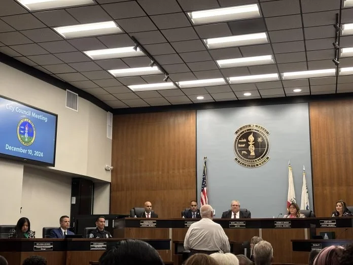 City council meeting in a formal chamber with six officials seated at a raised desk, a speaker standing at the podium addressing the council, flags behind the officials, and a large display screen showing the date December 10, 2024.