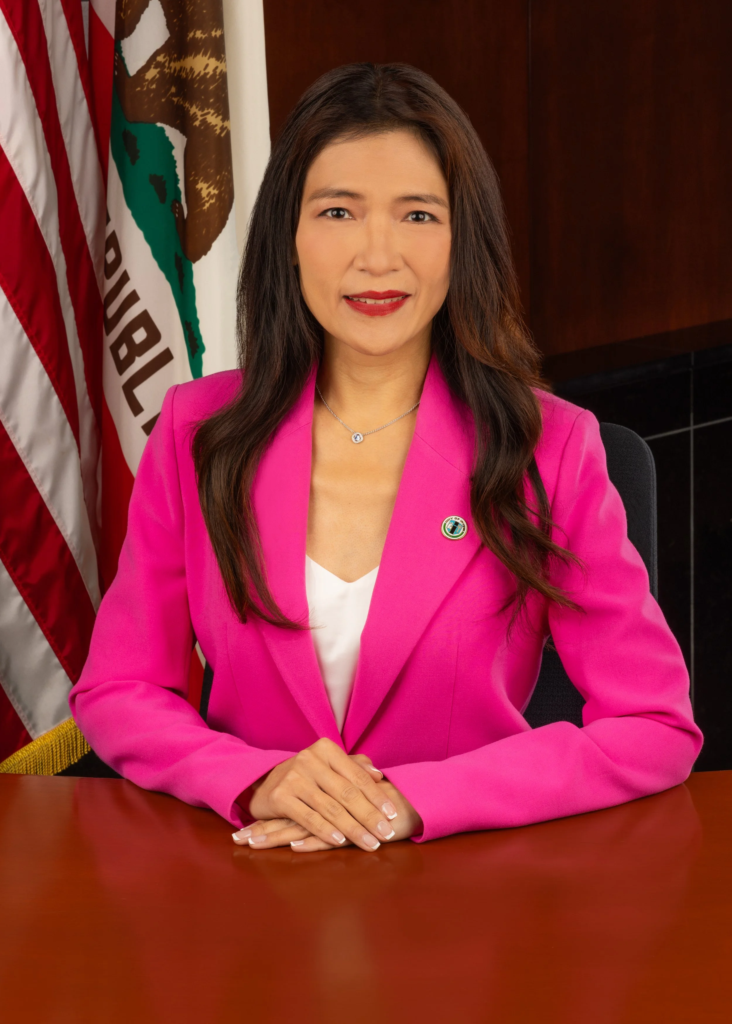 A woman in a bright pink blazer seated at a table with her hands clasped, smiling, with a California flag and a wooden wall in the background.