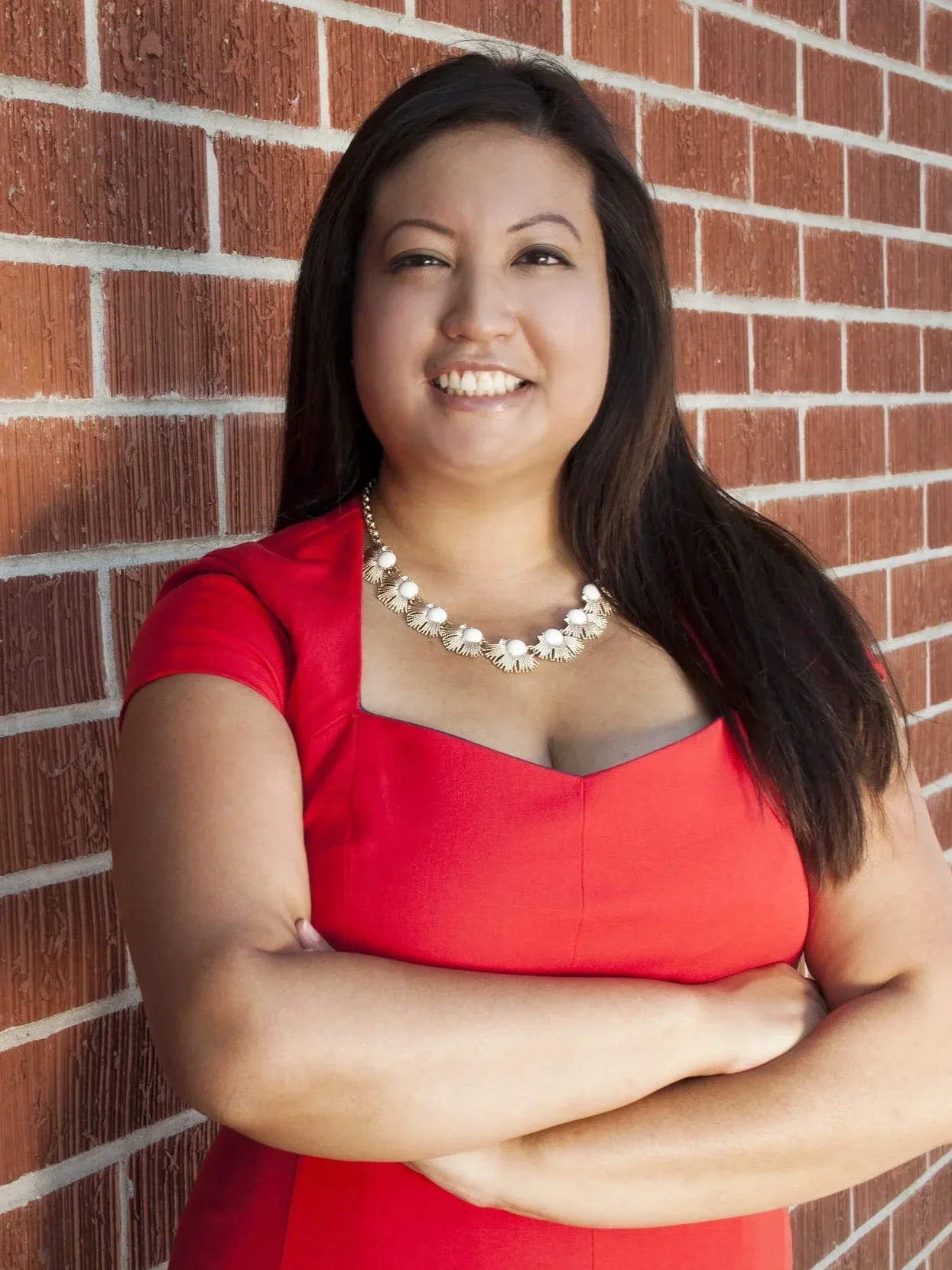 A woman with dark hair, wearing a red dress and a pearl necklace, standing in front of a brick wall, smiling with arms crossed.
