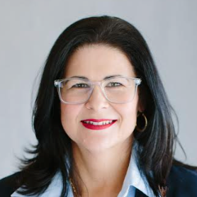 A woman with shoulder-length black hair, wearing glasses, gold earrings, and a light blue shirt, smiling against a light gray background.