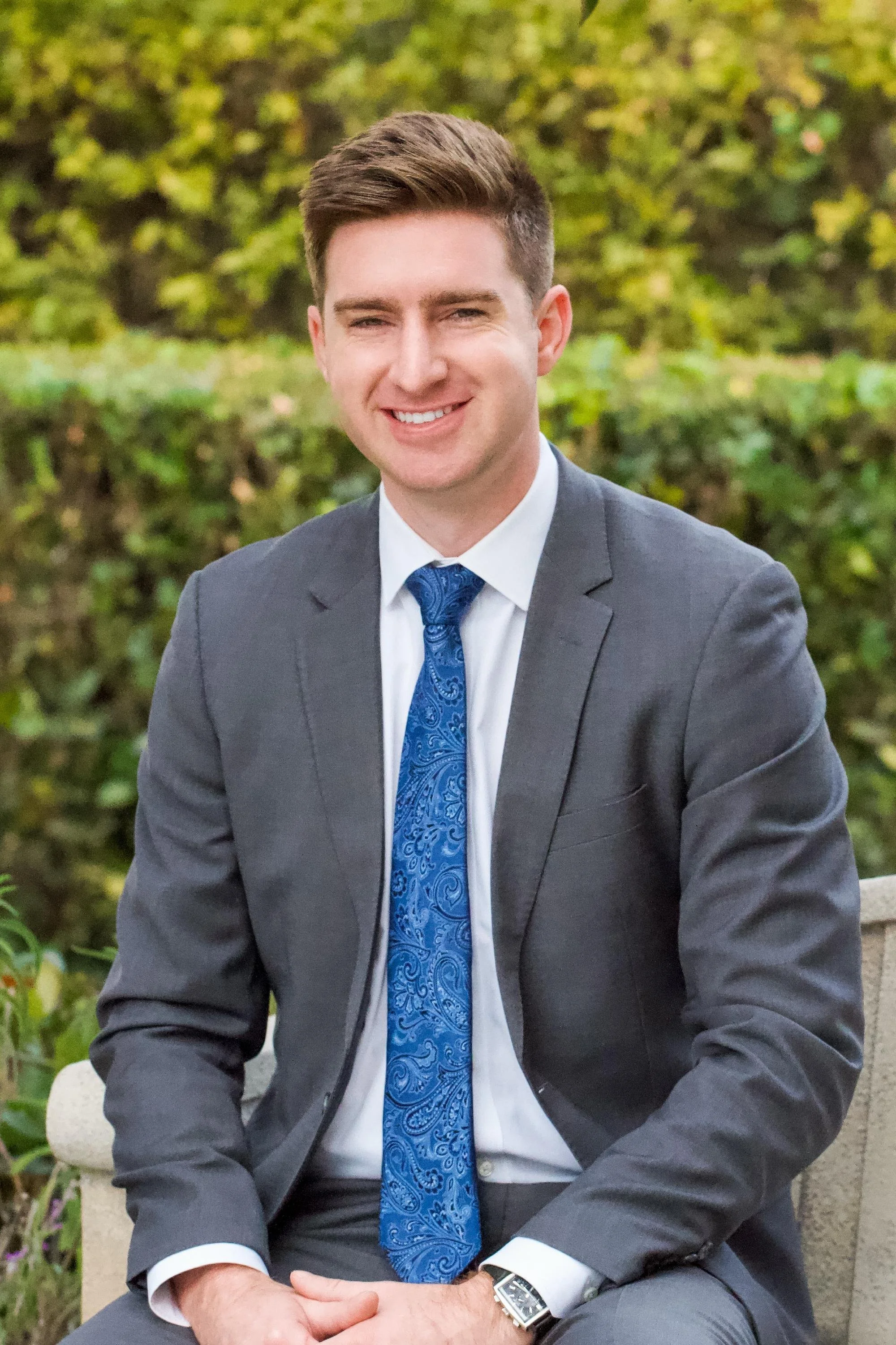 A young man in a gray suit with a blue patterned tie, white shirt, and a watch, sitting outdoors on a bench, smiling with a background of green bushes and trees.
