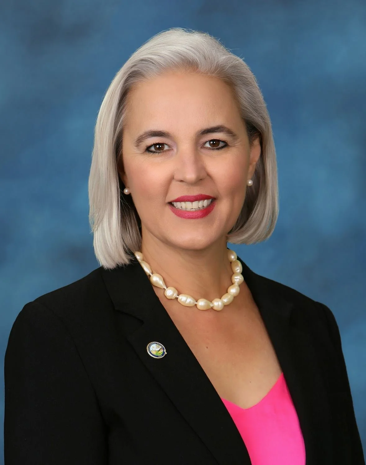 Portrait of a smiling middle-aged woman with gray hair, wearing a black blazer, pink top, pearl necklace, and pearl earrings, against a blue background.
