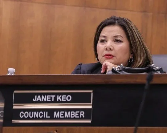 A woman sitting behind a nameplate that reads 'Janet Keo, Council Member' in a formal setting.