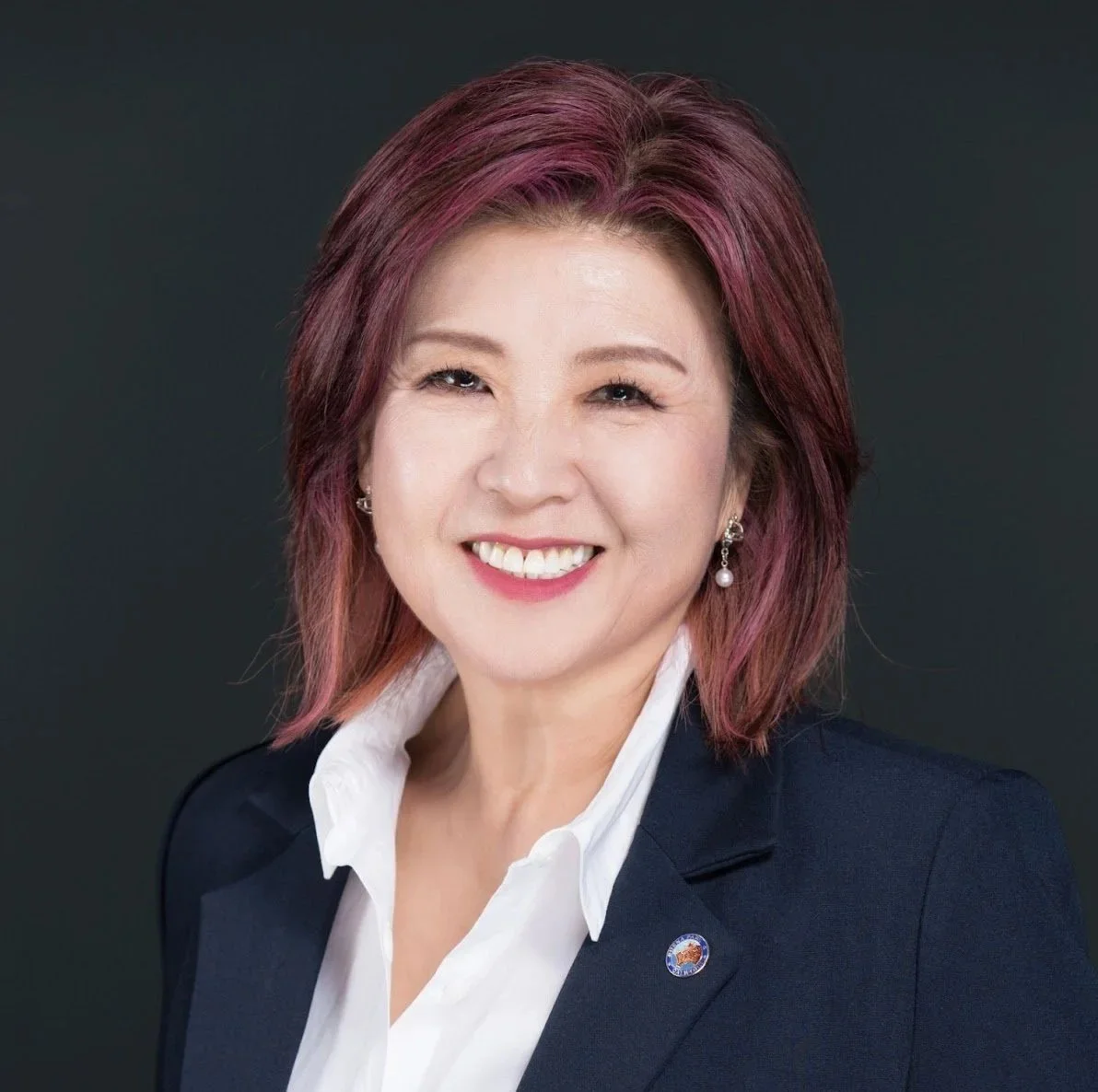 Portrait of a smiling woman with shoulder-length pinkish hair, wearing a navy blazer with a lapel pin and a white collared shirt, against a dark background.