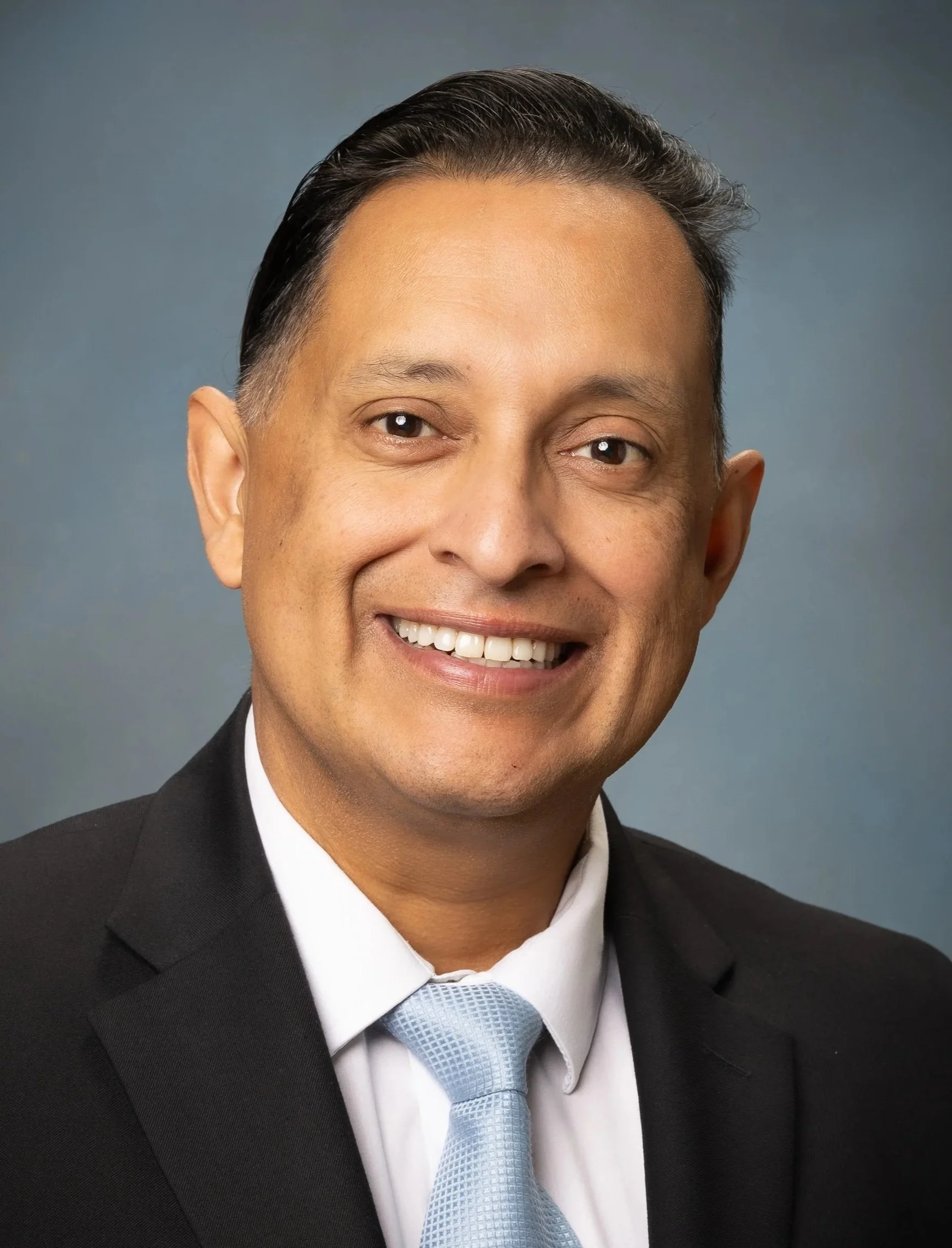 A professional headshot of a smiling man in a dark suit, white shirt, and light blue tie against a gray gradient background.