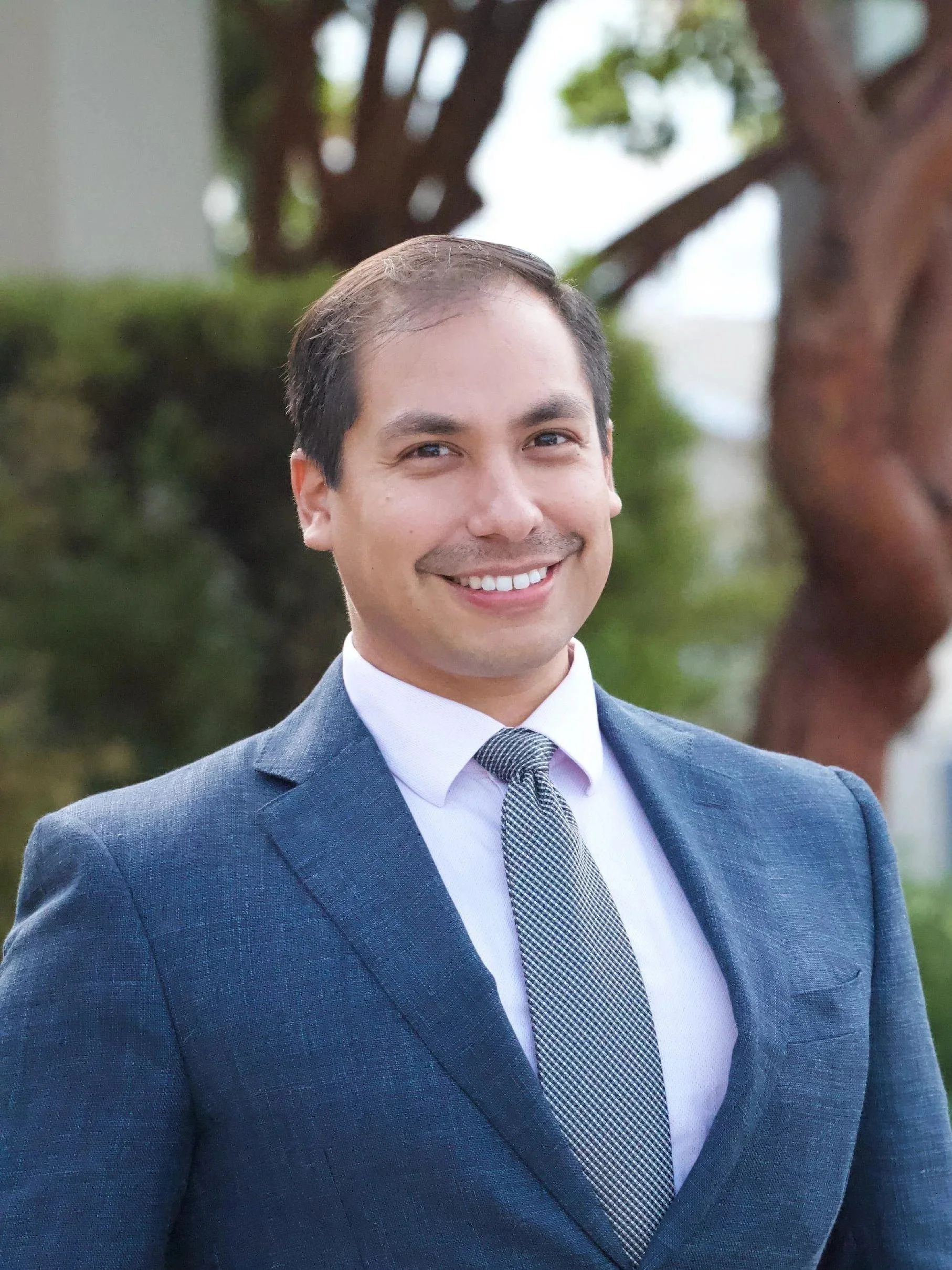 A man in a blue suit and patterned tie smiling outdoors with green trees in the background.