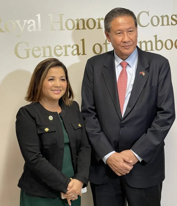 A woman and a man standing side by side in front of a beige wall with gold text. The woman has shoulder-length brown hair, is smiling, and is wearing a black blazer with gold buttons over a green dress. The man has short gray hair, is looking slightly to the side, and is dressed in a dark suit with a light blue shirt and a pink tie. The background text includes parts of words like 'Royal Honor,' 'General,' and 'Cons'.