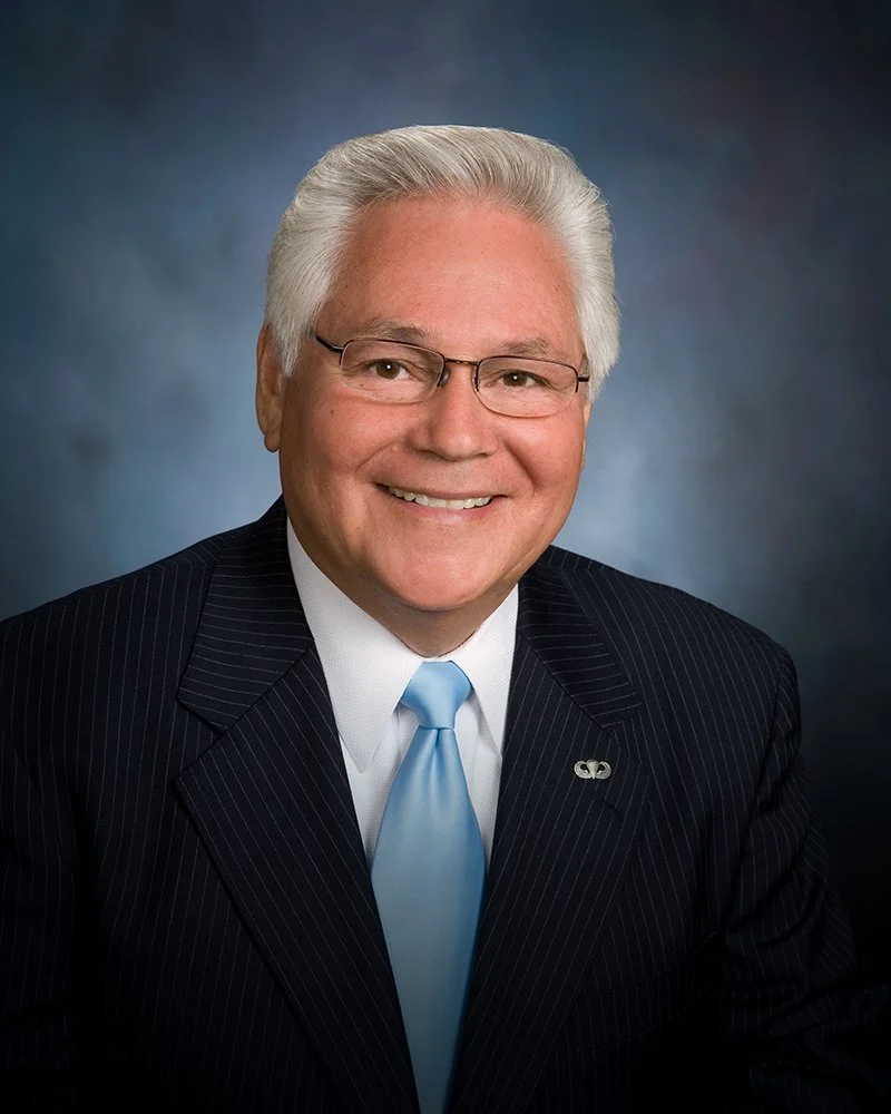 A smiling older man with gray hair and glasses, dressed in a dark pinstripe suit, white shirt, and light blue tie, against a dark blue background.