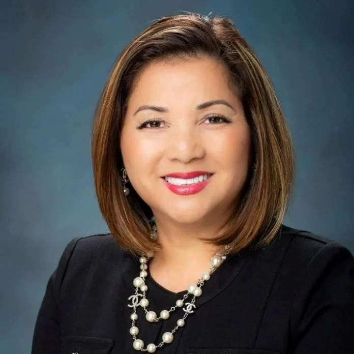 A woman with shoulder-length brown hair smiling, wearing a black blazer, pearl necklace, and earrings, against a blue gradient background.