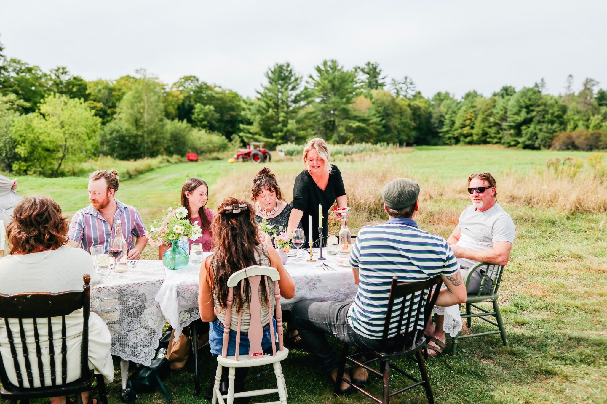 Group of people sitting around a table outdoors in a field, enjoying a meal on a cloudy day, with trees and a tractor in the background.