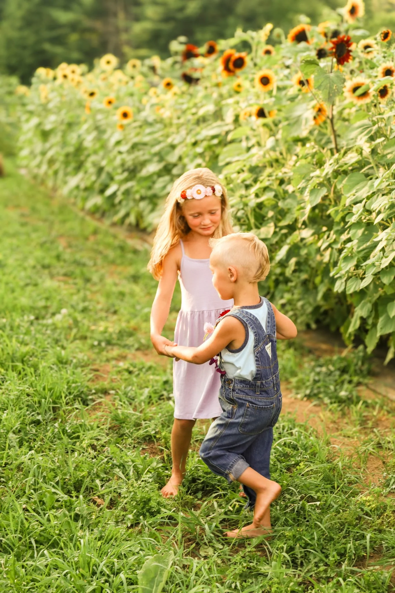 Two young children, a girl and a boy, are playing together in a sunflower field during daytime. The girl is wearing a light purple dress with a flower crown, and the boy is dressed in denim overalls. They are holding hands and appear to be happy.