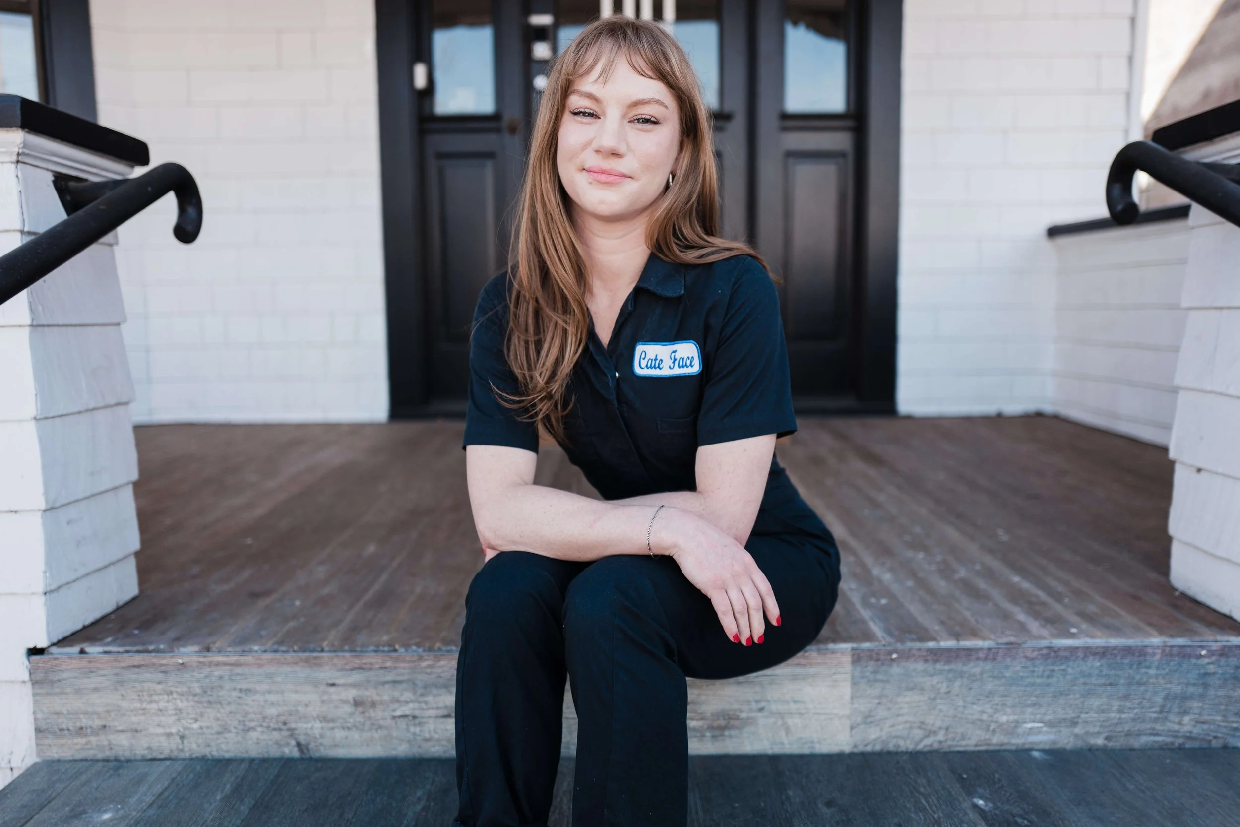 Cate Draney, Portland Esthetician, is sitting on the front steps of the Heal House, wearing a navy blue uniform with a name patch that reads 'Cate Face'.