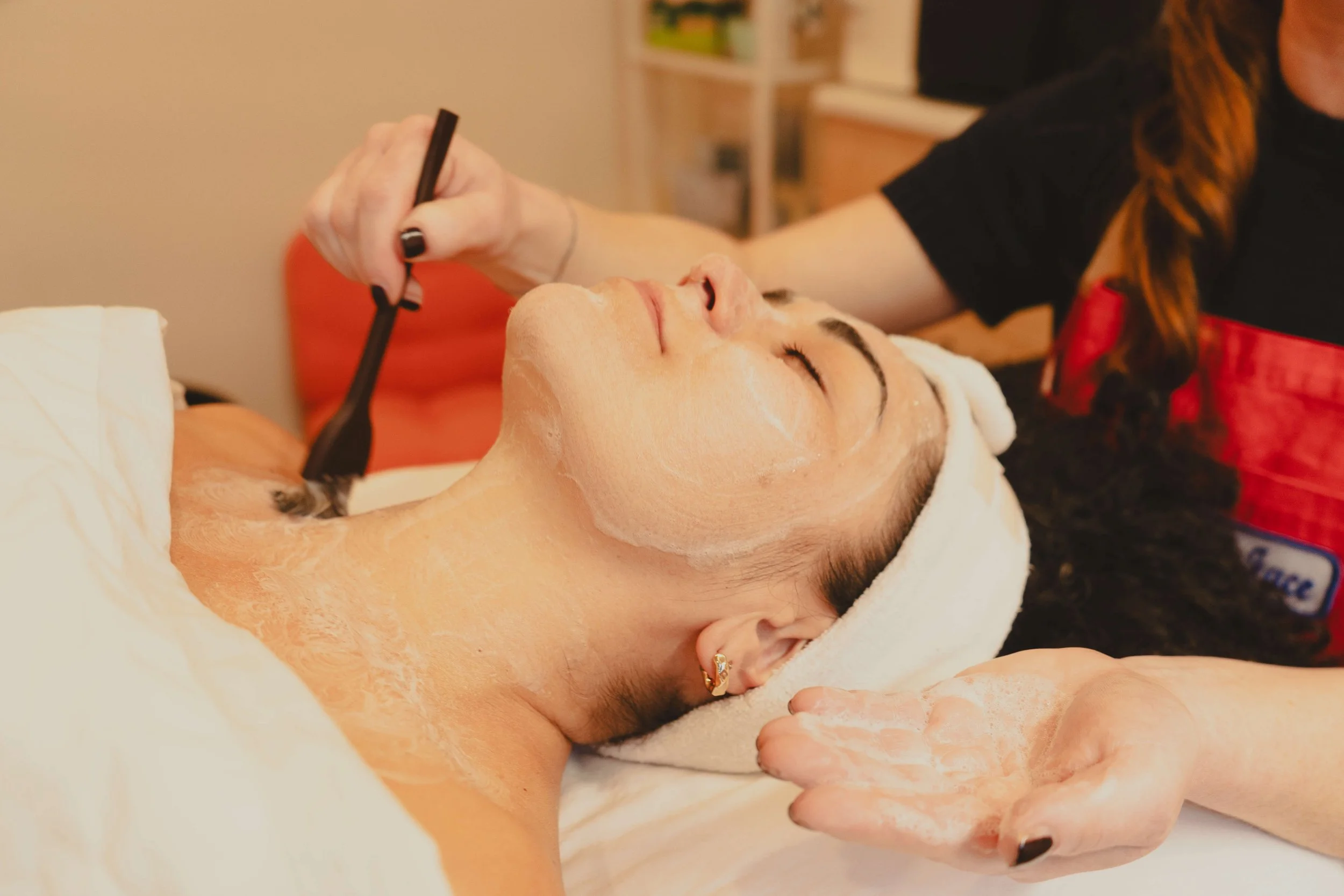 A woman receiving a facial treatment at a spa, lying down with a towel wrapped around her head, while Cate applies a mask on her face.
