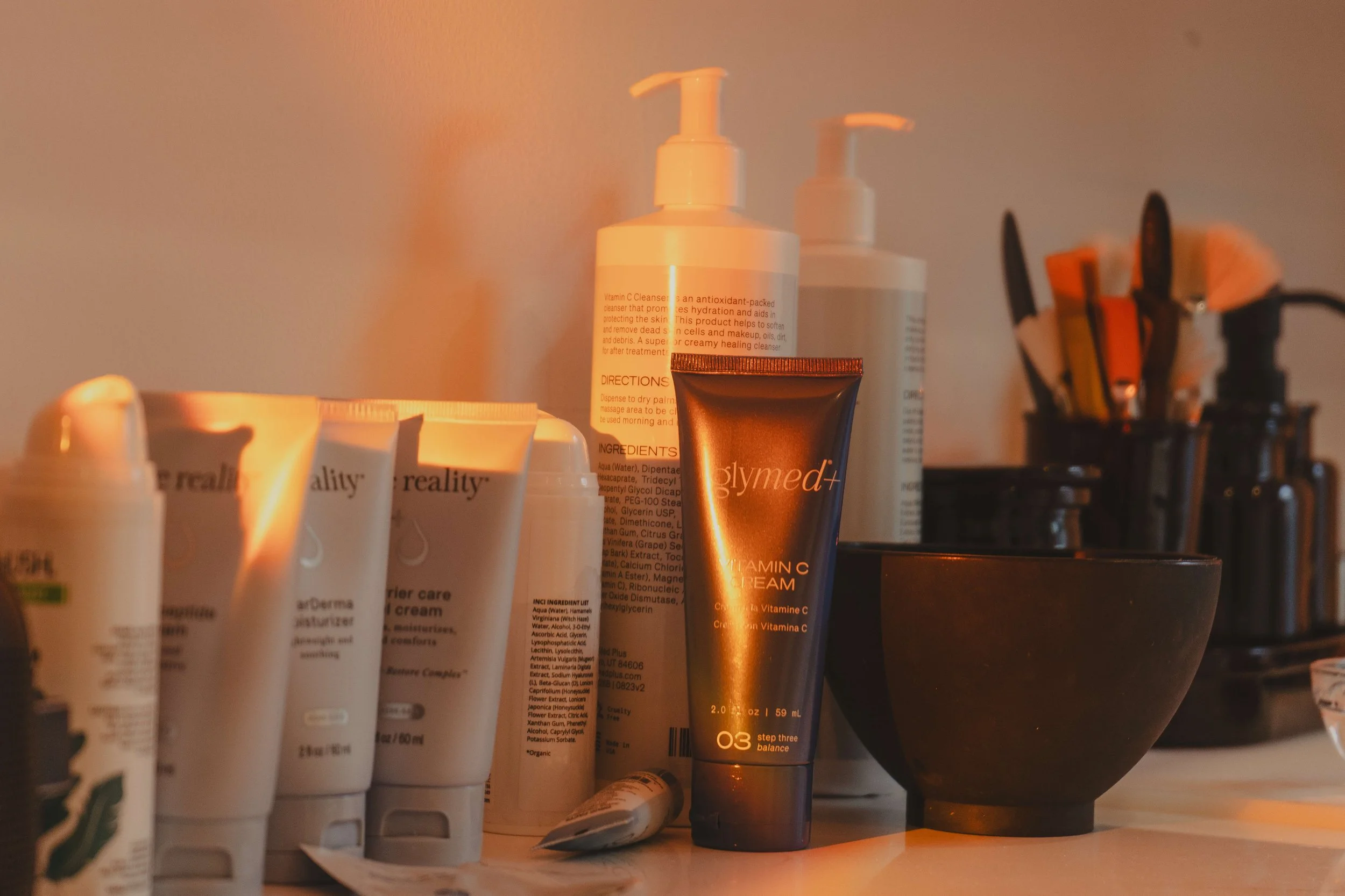 Various skincare and beauty products on a bathroom countertop, including tubes, bottles, a black bowl, and brushes in a container, with warm lighting.