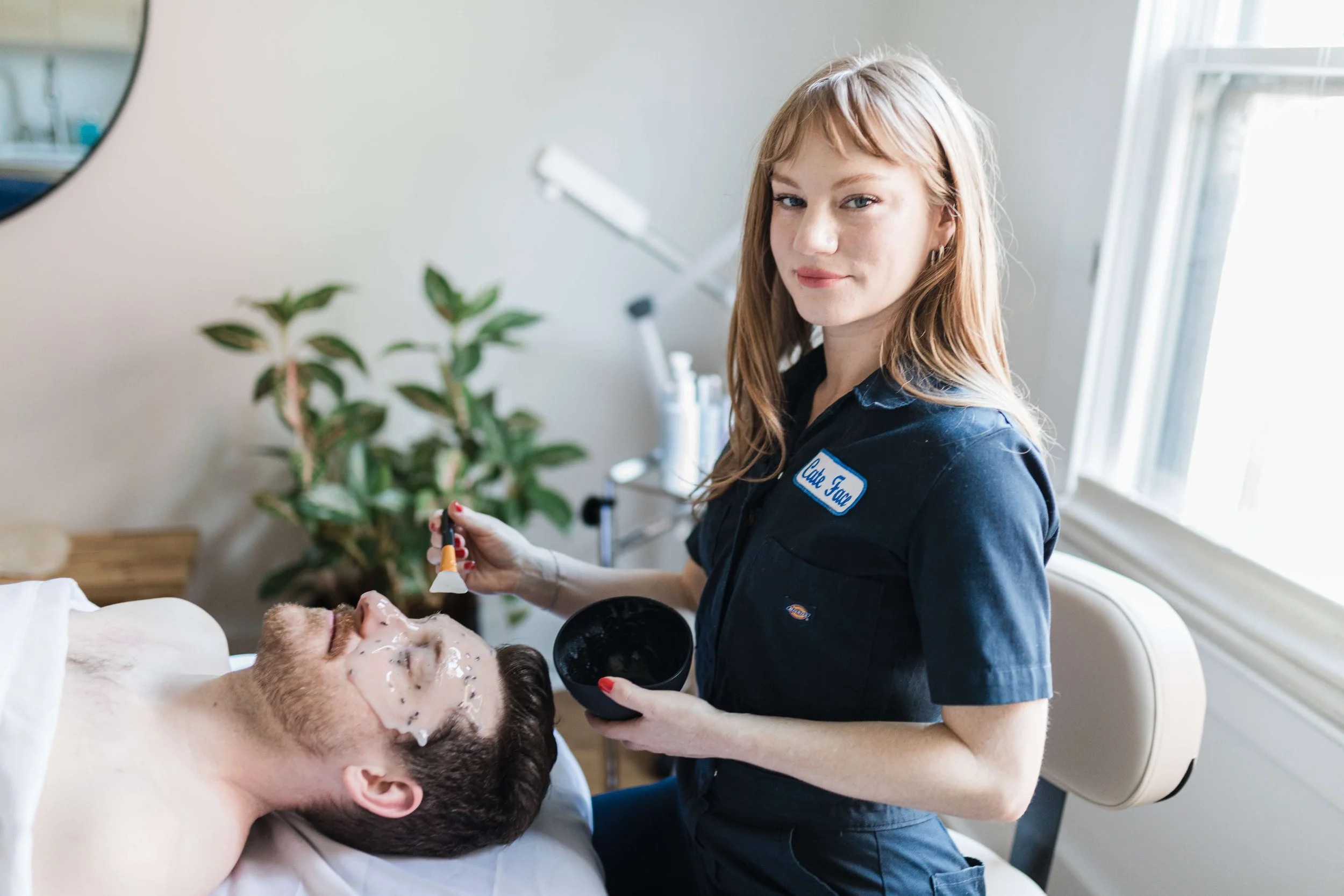 Cate Draney, Portland Esthetician, is applying a facial mask to a man's face in her private treatment room, with a green plant and skincare products in the background.
