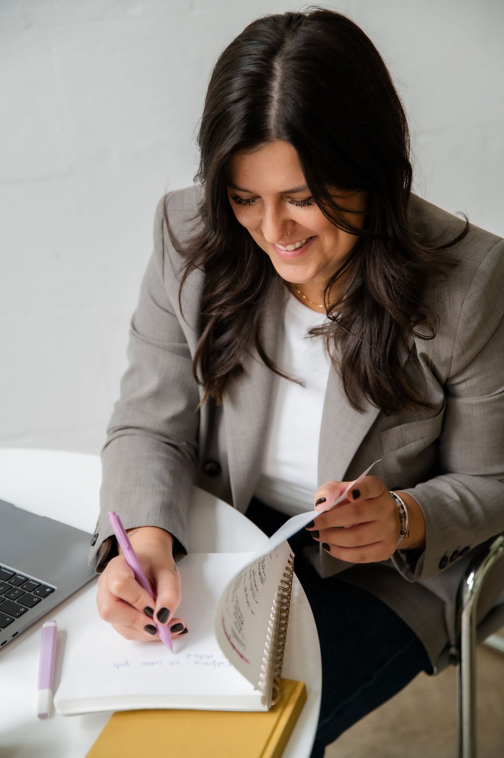 A woman with long dark hair, dressed in a gray blazer and white top, is sitting at a table writing in a notebook with a pink pen. She is smiling and appears to be working or studying.