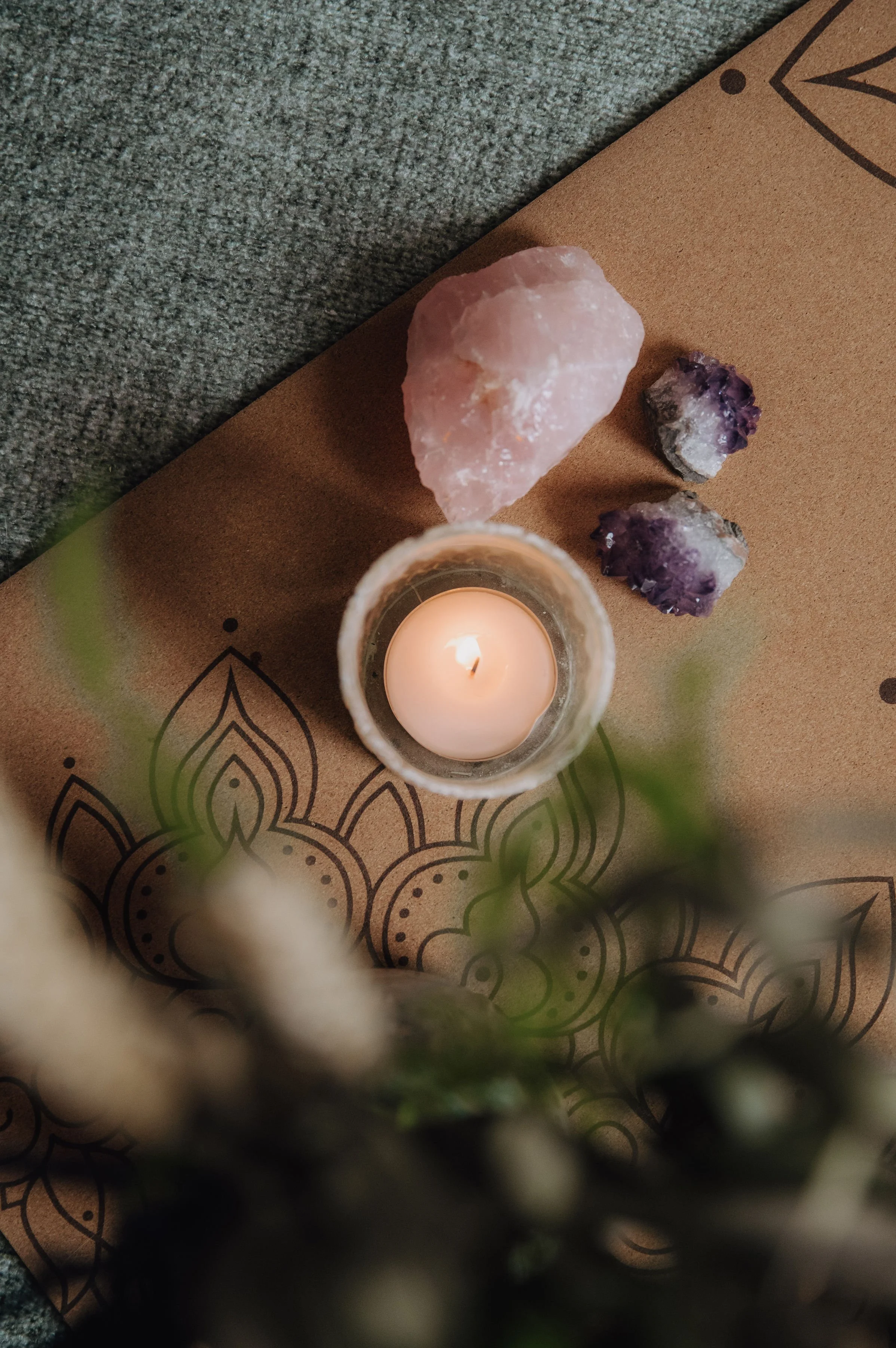 A lit candle in a glass holder, surrounded by three large crystals (pink and purple), placed on a piece of brown paper with black mandala designs, and a gray textured surface underneath.