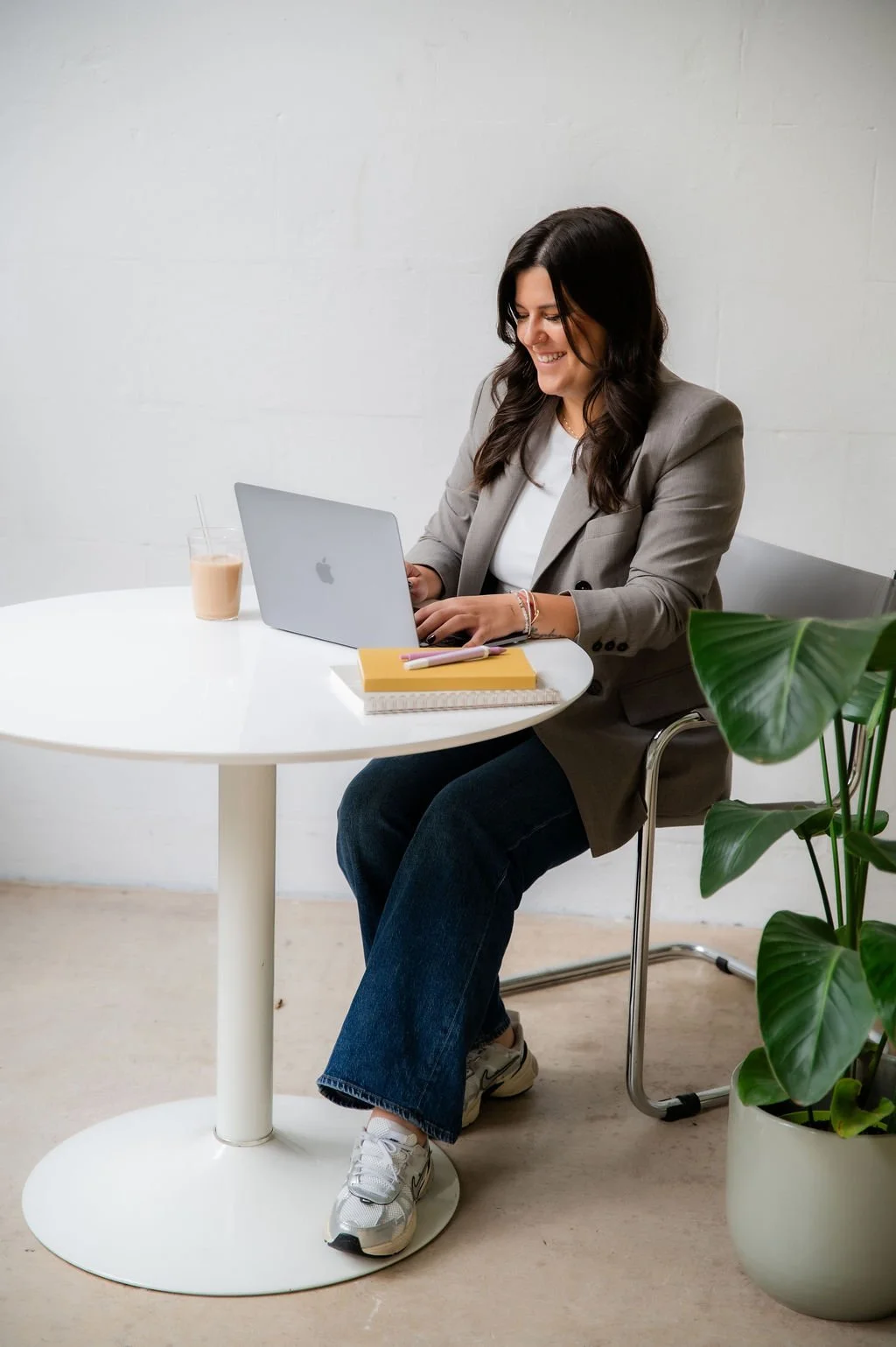A woman sits at a round white table working on a silver laptop, with a yellow notebook, a spiral notebook, a glass of iced coffee, and a straw, and a large green potted plant nearby.