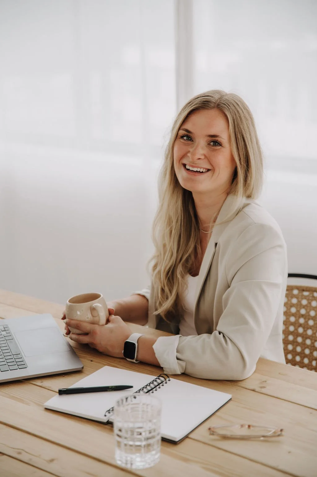 A woman with long blonde hair sitting at a wooden desk, smiling, holding a beige coffee mug, with a laptop, notebook, pen, glass of water, and glasses on the desk.
