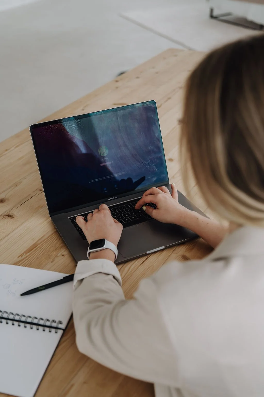 Person using a MacBook Pro on a wooden table, with a notebook and pen nearby.