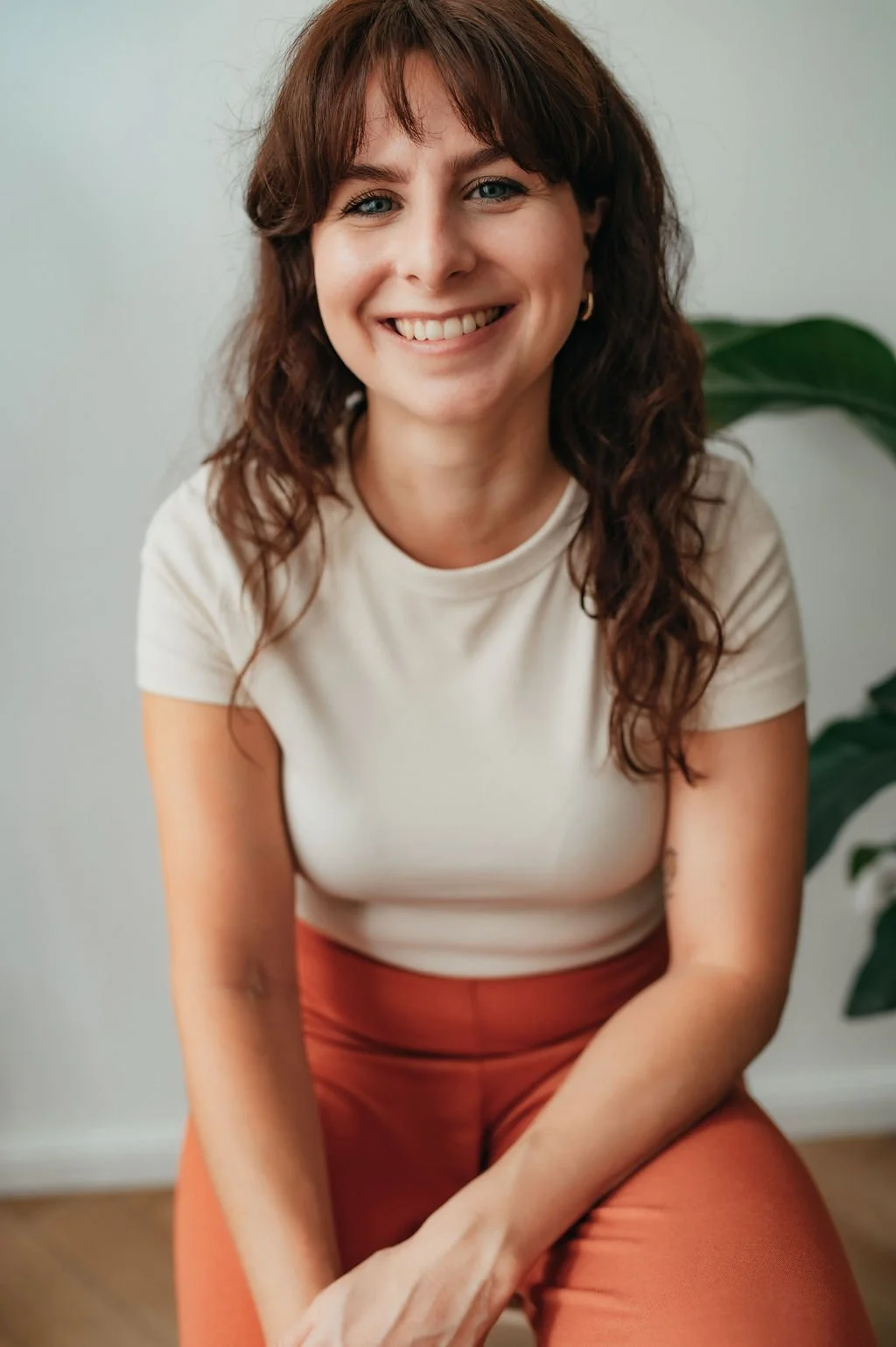 A woman with shoulder-length brown hair and blue eyes, smiling, wearing a white t-shirt and rust-colored pants, sitting indoors near a green plant.