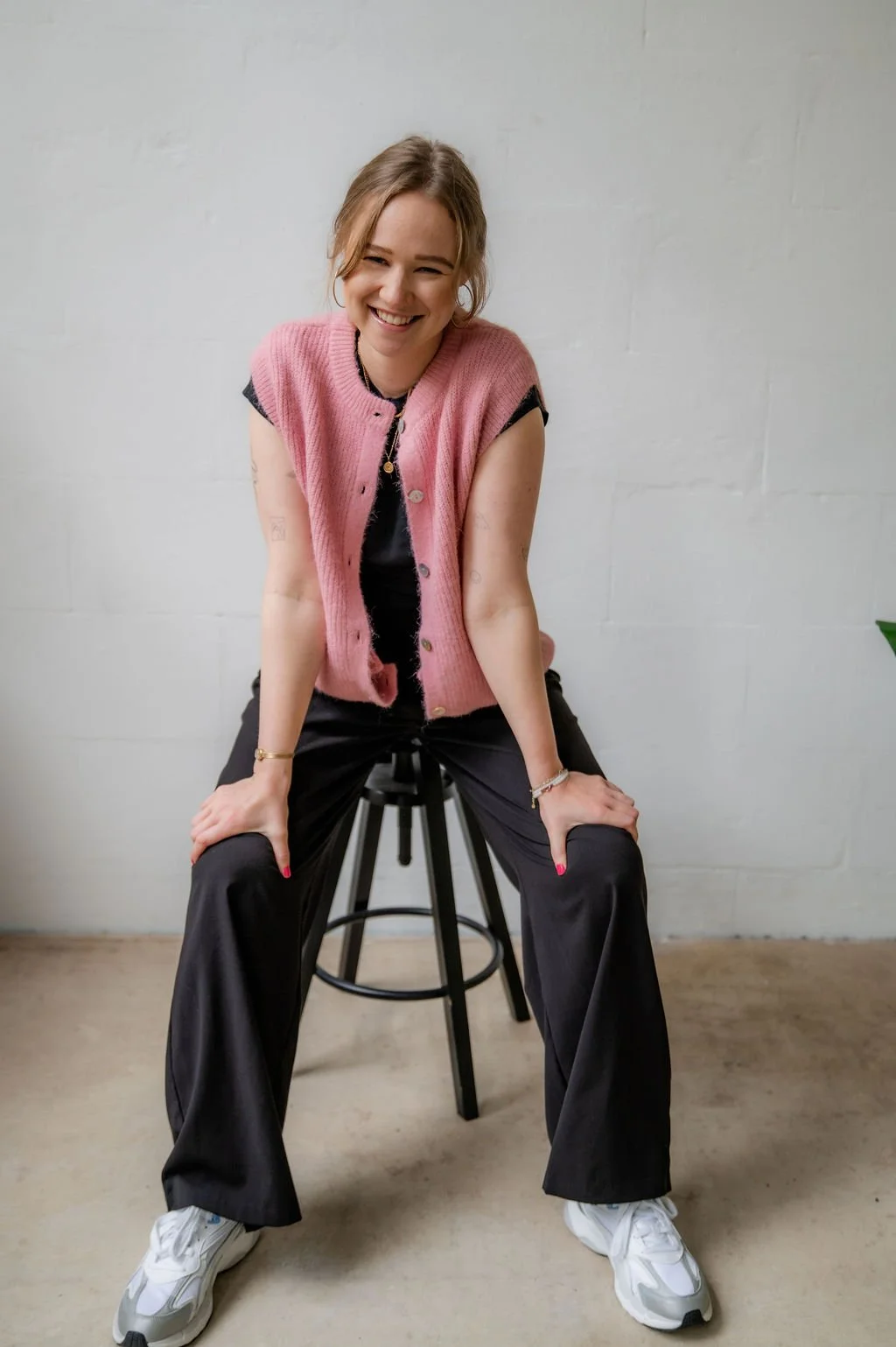A woman with light brown hair, wearing a pink vest over a black top and black wide-leg pants, sitting on a black stool against a plain white wall, smiling at the camera.