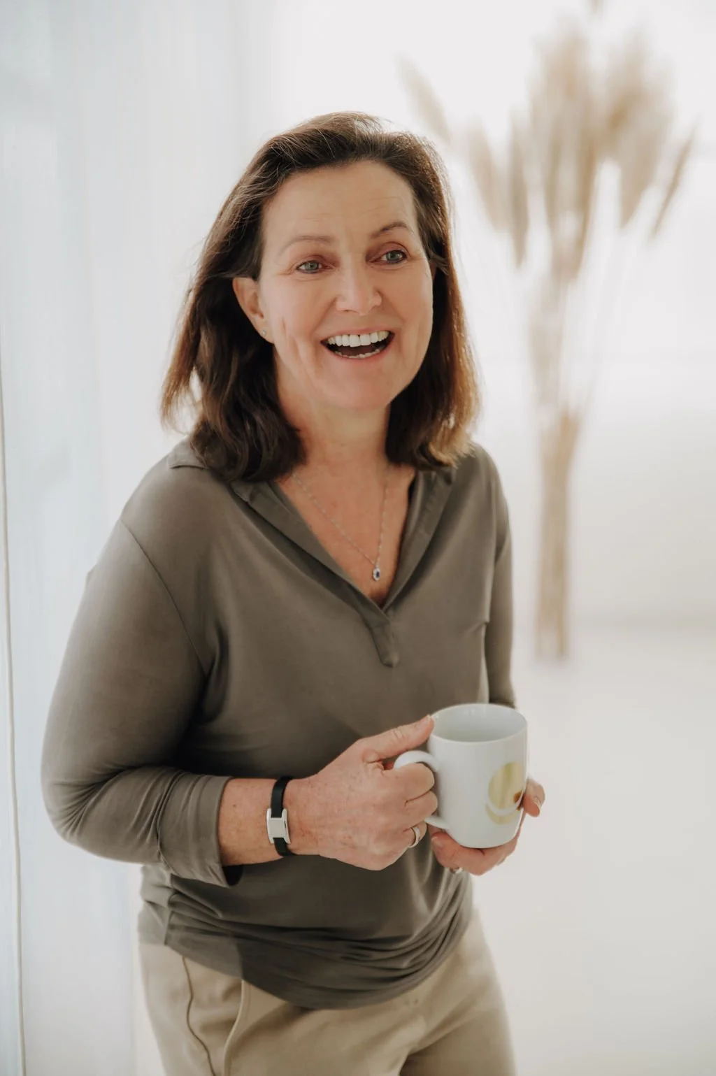 A smiling woman with shoulder-length brown hair holding a white ceramic mug in a bright, minimalist room.