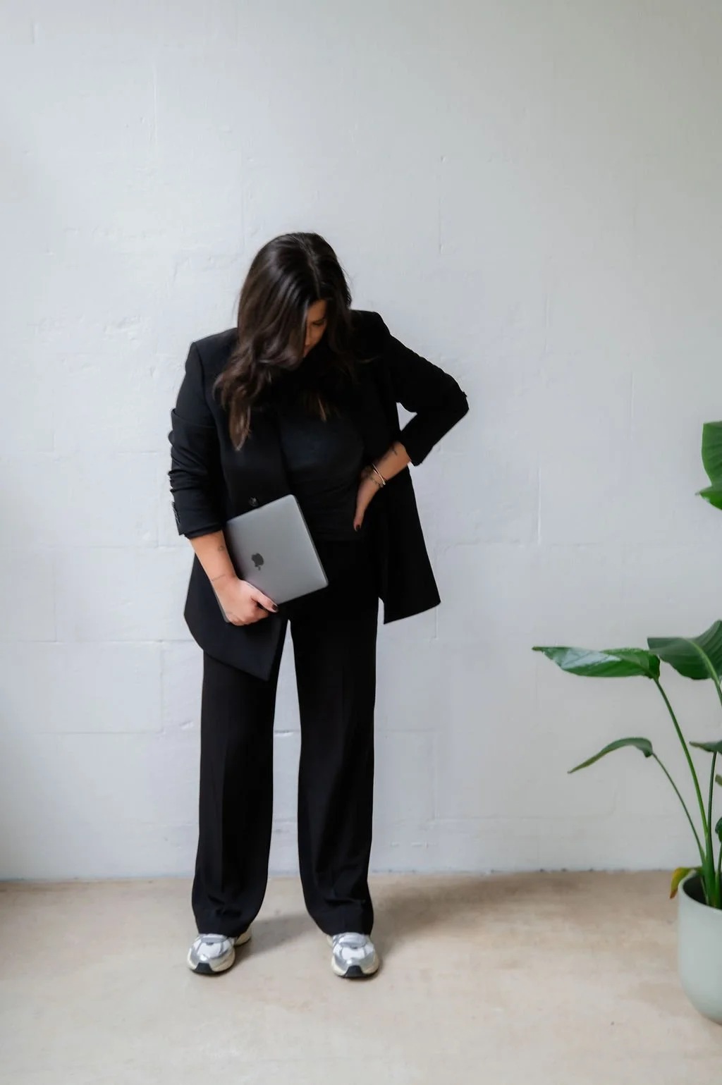 A woman dressed in black, holding a MacBook, looking down, standing against a plain white wall with a large green plant on the right.