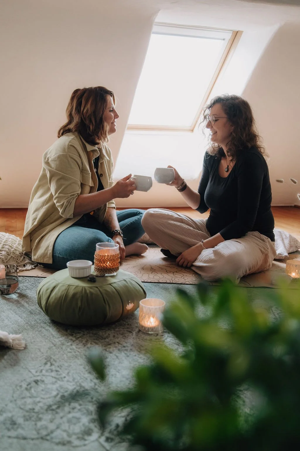 Two women sitting on a rug on the floor, sharing drinks and smiling at each other in a cozy, well-lit attic room with a skylight.