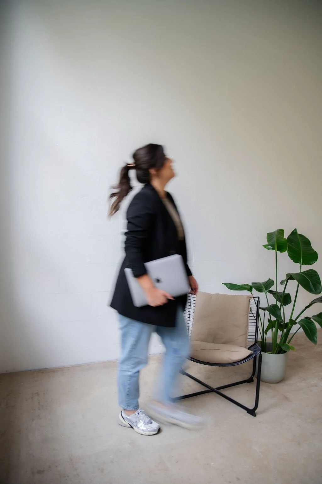 A woman walking indoors, holding a silver laptop, wearing a black blazer, jeans, and sneakers, with a large green plant and a beige chair in the background.