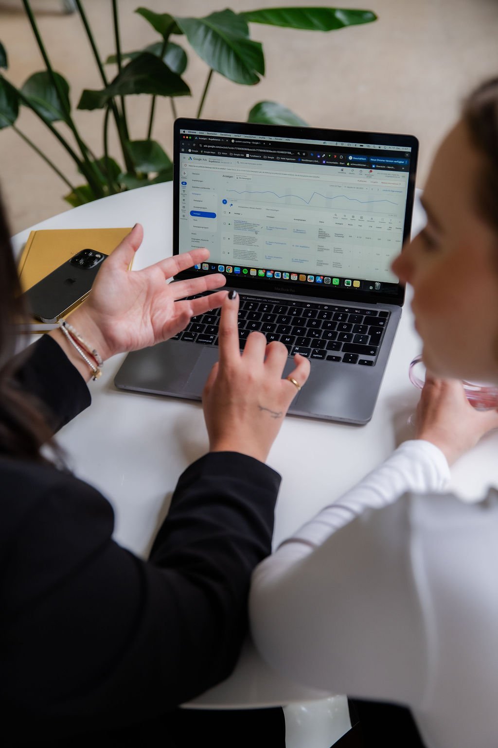 Two women working on a laptop at a white table, discussing data with a Google Analytics dashboard visible on the screen. One woman is pointing to the screen while the other watches.