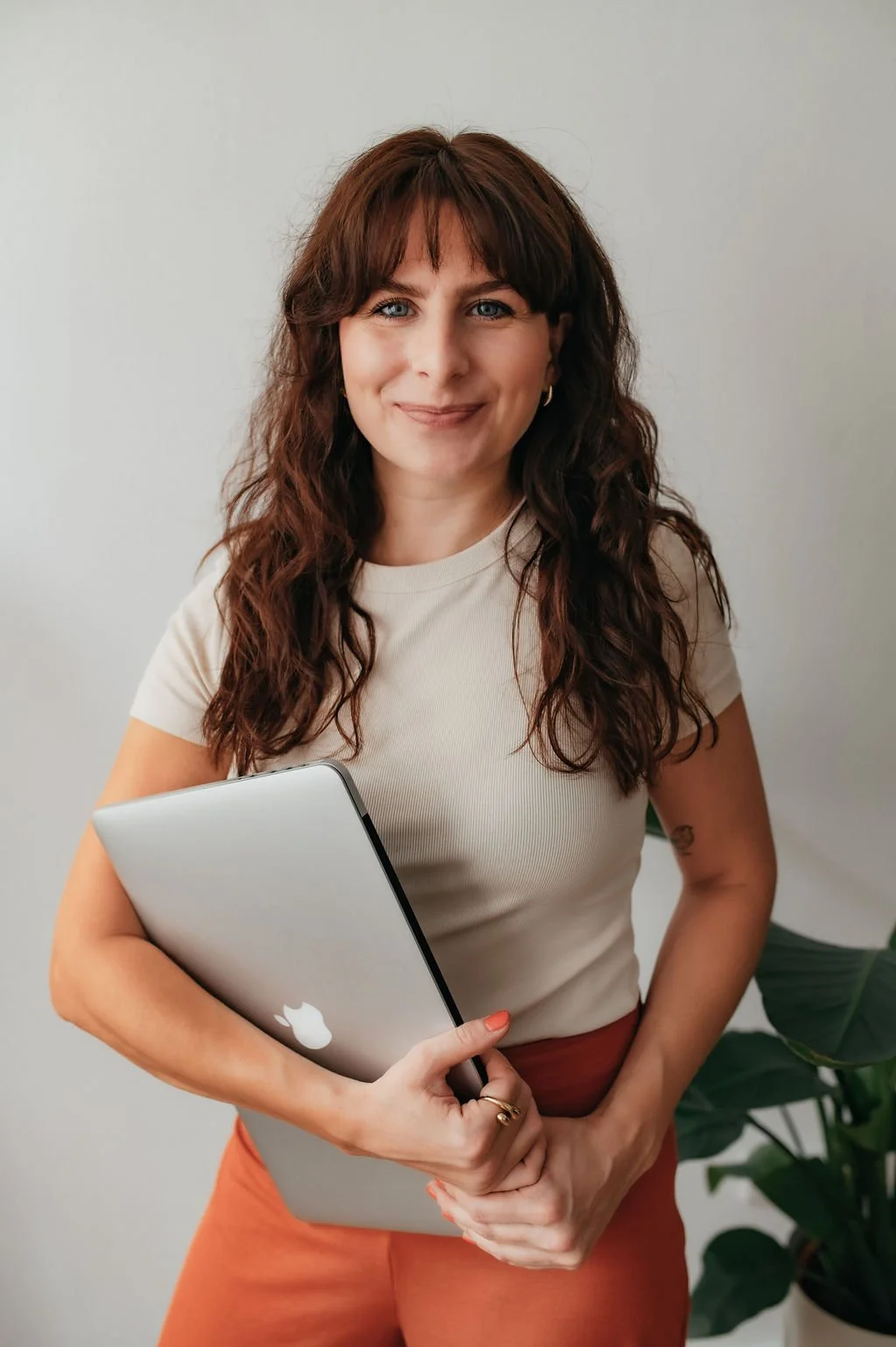 A woman with wavy brown hair, wearing a white t-shirt and rust-colored pants, holding a closed MacBook laptop, smiling, standing next to a green plant against a plain light background.