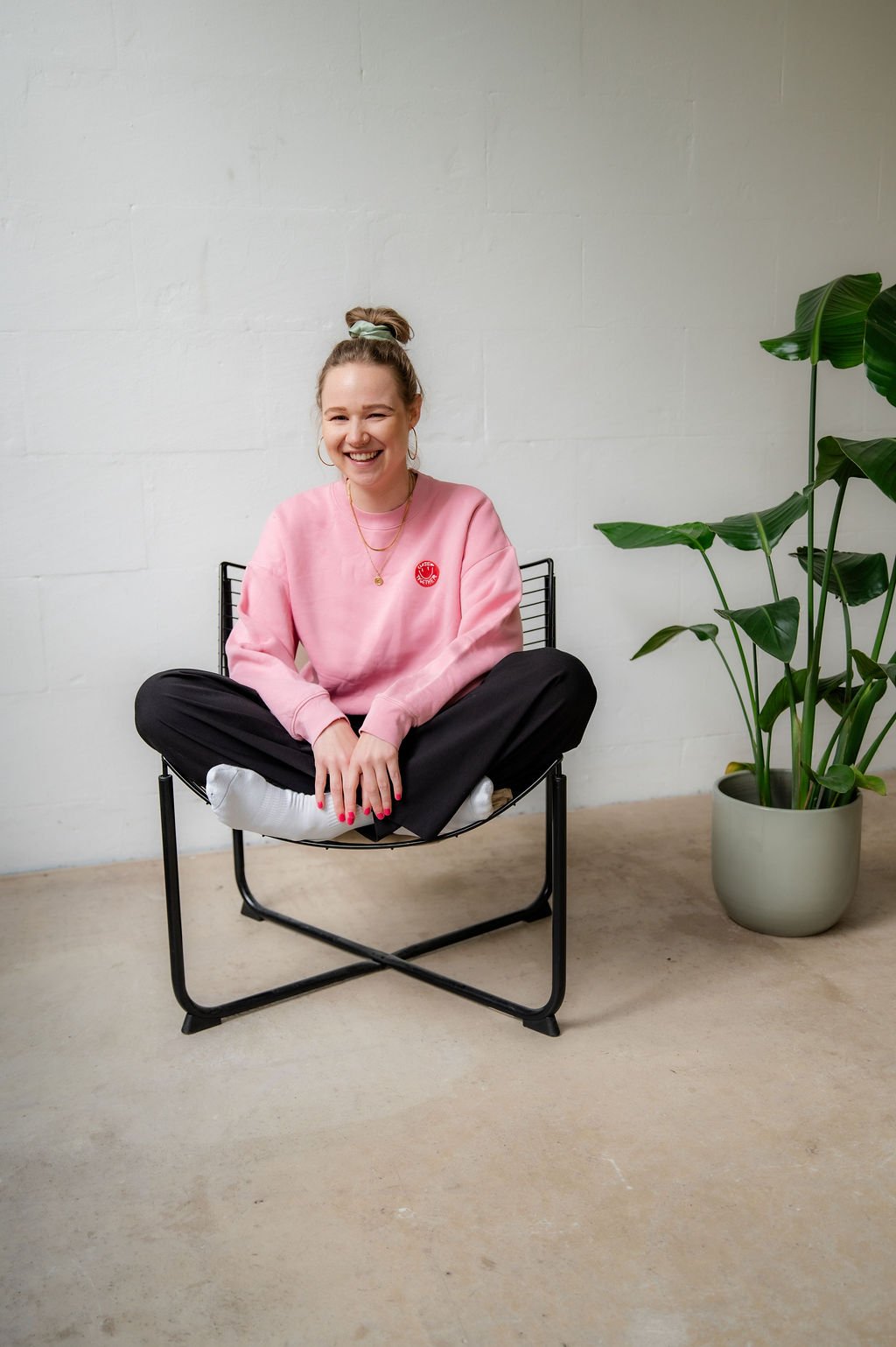 A woman with a top bun hairstyle, wearing a pink sweatshirt, black pants, white socks, and jewelry, sitting cross-legged on a black wire chair, smiling, with a large green potted plant beside her against a plain white wall.