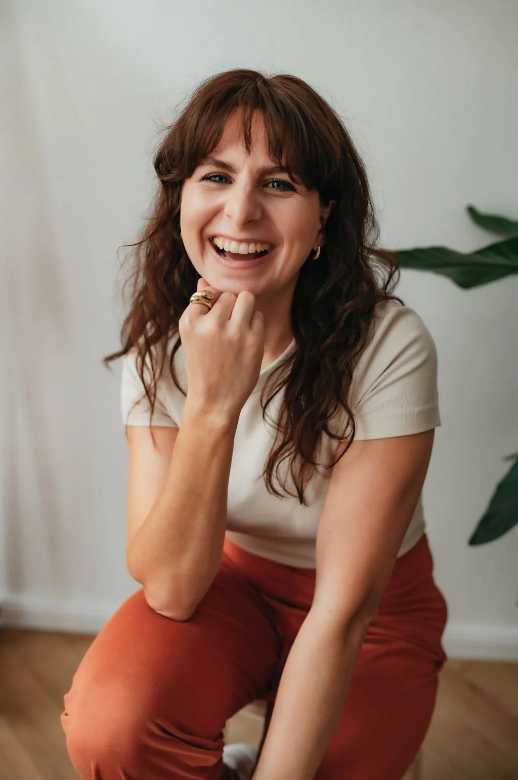 A woman with curly brown hair smiling, wearing a beige top and rust-colored pants, sitting indoors next to a green plant.