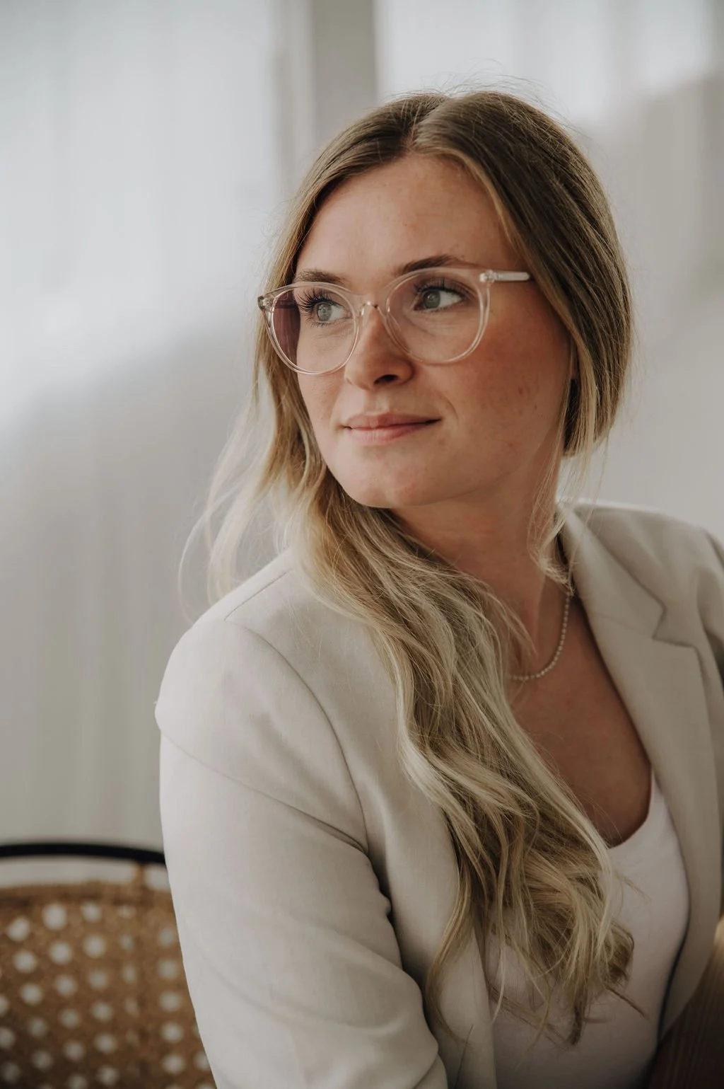 A young woman with long, wavy blonde hair, wearing clear glasses and a white blouse, looking to her right with a subtle smile.